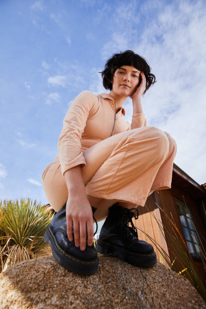 Stylish young woman crouching outside on a rock Portrait of a stylish young woman in a jumpsuit and platform boots crouching on a rock outside against a blue sky squatting position photos stock pictures, royalty-free photos & images