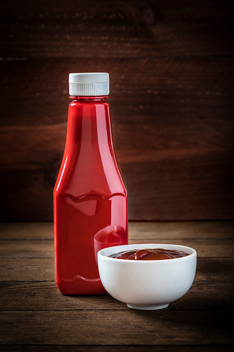 Front view of a ketchup bottle alongside a white bowl full of ketchup sauce on a dark rustic wooden table. Low key DSLR photo taken with Canon EOS 6D Mark II and Canon EF 24-105 mm f/4L