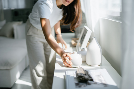 Young Asian woman enjoys her time at home, decorating and organising picture frames and vases on the shelf by the window
