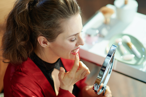 young woman in black body lingerie and red bathrobe in the modern house in sunny day looking in mirror and checking teeth.