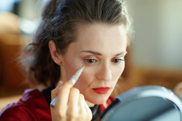 woman looking in mirror and applying liquid eye concealer stylish woman in black body lingerie and red bathrobe in the modern living room in sunny day looking in mirror and applying liquid eye concealer. concealer stock pictures, royalty-free photos & images