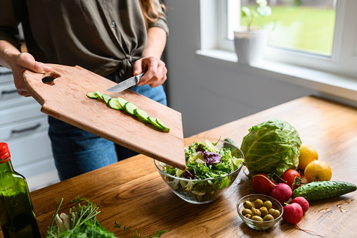Cooking salad at home. A woman throws cucumber from cutting board in salad, fresh vegetables and greens on the table near. Healthy food concept. Face is not visible