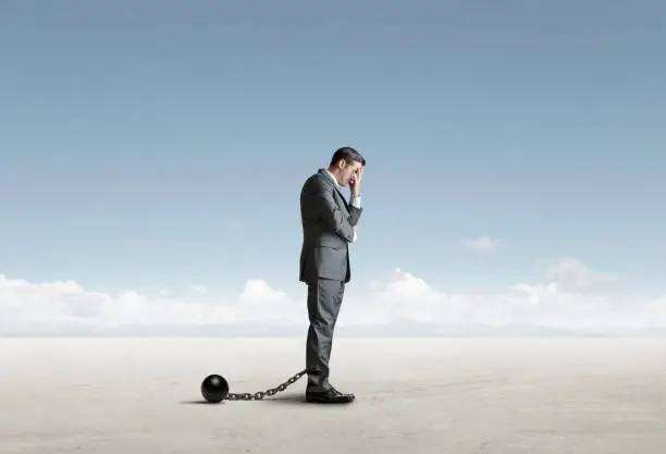 A ball and chain is attached to a businessman's leg as he places his head in his hands in a sign of resignation.