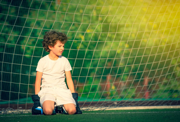 nachdenklicher kleiner junge sitzt auf dem grünen rasen in den torpfosten auf einem sportplatz young goalie. junge torwart in fußball-sportbekleidung auf stadion mit ball. sport concept.copy space - playing field goalie soccer player little boys stock-fotos und bilder