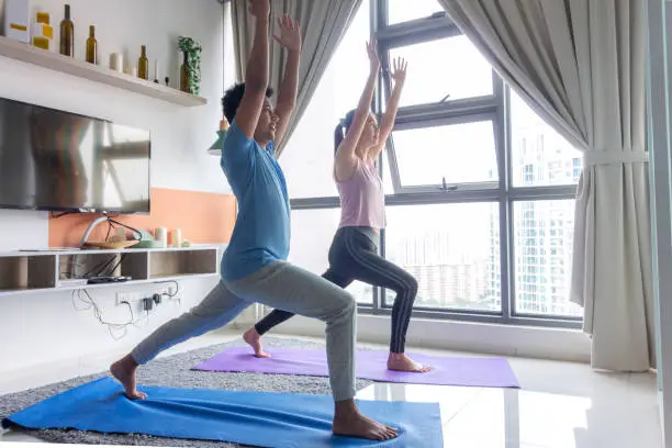 Asian male and female partners are practicing yoga warrior pose, Virabhadrasana in apartment during pandemic lockdown