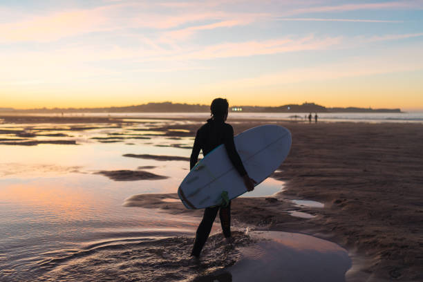 Young male surfer walk on beach sand at sunset with board under his arm