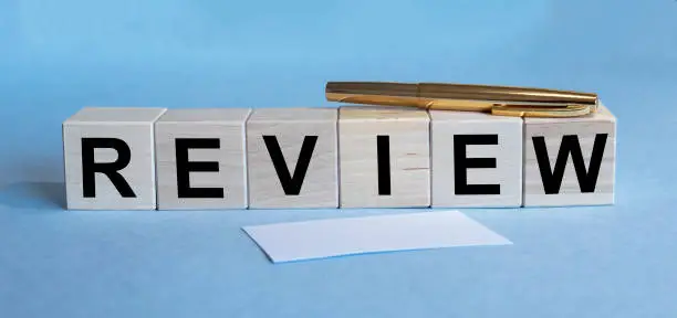 Photo of Word REVIEW written on wooden cubes. Pen and paper and cubes on blue table