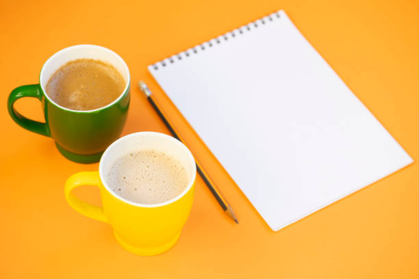 Two cup of cappuccino, paper notepad with pencil on the orange background. Empty space for text.
