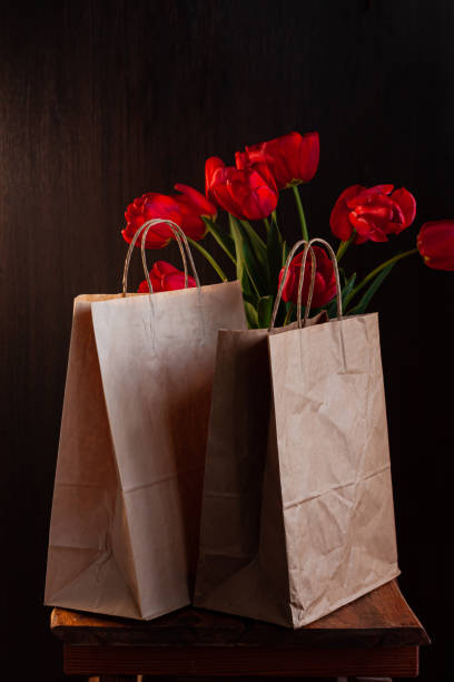 Kraft paper bags with blooming red tulips bouquet on black background.Donations help recycling boxes, dark moody low key Kraft paper bags with blooming red tulips bouquet on black background. Dark moody low key minimalism style flowers and food delivery packaging banner copy space mockup. Donations help recycling boxes. Slective Focus stock pictures, royalty-free photos & images