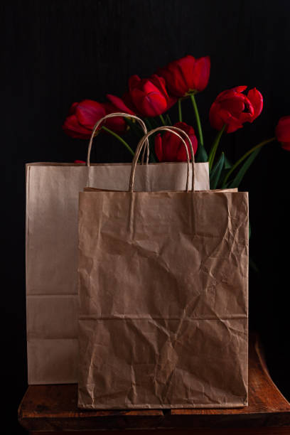 Kraft paper bags with blooming red tulips bouquet on black background.Donations help recycling boxes, dark moody low key Kraft paper bags with blooming red tulips bouquet on black background. Dark moody low key minimalism style flowers and food delivery packaging banner copy space mockup. Donations help recycling boxes. Slective Focus stock pictures, royalty-free photos & images