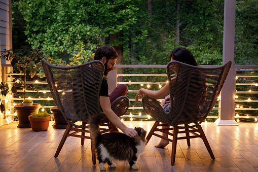 A couple sit in chairs and hold hands on a home patio.  It is early evening, and some string lights on the patio railing are on.  They are facing away from the camera looking out into some trees beyond the patio.  The man is petting a cat that is beside them.  They are relaxing together after a long day.  Shot during the covid-19 related isolation, this is the only place they can go out to relax.  The man is caucasian, the woman is Iranian ethnicity.