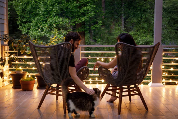 Couple on Patio in the Evening A couple sit in chairs and hold hands on a home patio.  It is early evening, and some string lights on the patio railing are on.  They are facing away from the camera looking out into some trees beyond the patio.  The man is petting a cat that is beside them.  They are relaxing together after a long day.  Shot during the covid-19 related isolation, this is the only place they can go out to relax.  The man is caucasian, the woman is Iranian ethnicity. porch stock pictures, royalty-free photos & images