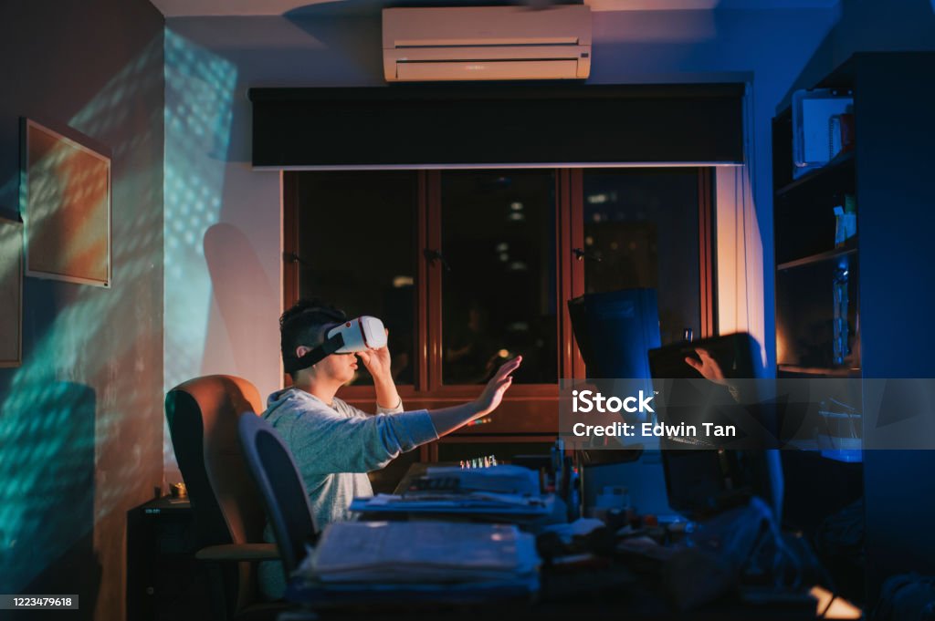 an asian chinese male put on VR goggle and experiencing 3D virtual gaming experience in his home office study room at night in front of his desktop PC Video Game Stock Photo