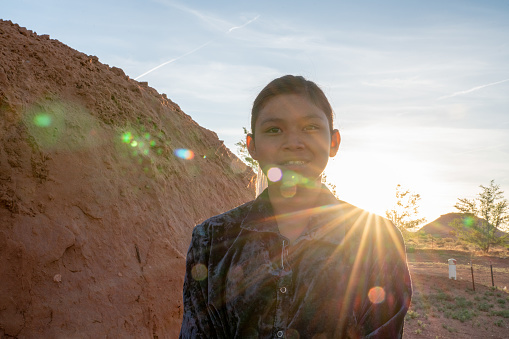 A happy teenage Navajo girl not wearing a mask for protection from Covid19 as she stands by a hogan at sunset