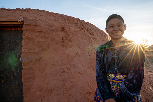 A happy teenage Navajo girl not wearing a mask for protection from Covid19 as she stands by a hogan at sunset