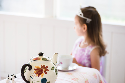 The young girl, dressed up as a princess, sits by herself in her room,  She is pretending to have tea.