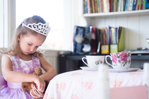 The young girl helps her toy bear dress up for tea by putting bracelets on his arm.  She is dressed as a princess.