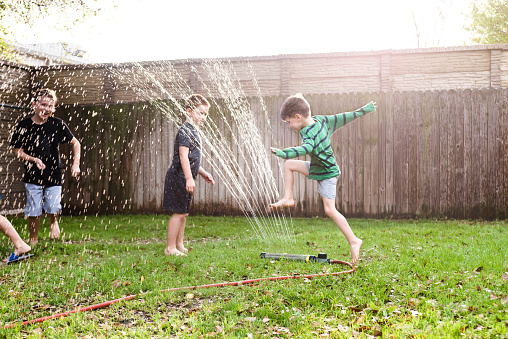 Brothers run and jump over the sprinkler to cool off on a hot summer day.  They are playing in their backyard.