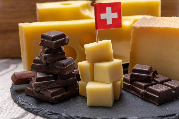 nourriture suisse savoureuse, bloc de fromage jaune moyen-dur emmental ou emmentaler avec des trous ronds, gruyère mûri et chocolat au lait de haute qualité servi en cubes comme haut de montagne avec drapeau suisse. - culture suisse photos et images de collection