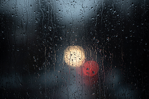 Looking through a window at a street with car's front and back light on a rainy and stormy late afternoon, focus on raindrops creating a bokeh