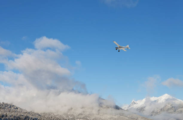 Small airplane flying over Southeast Alaskan mountains in winter with low clouds on a sunny day.