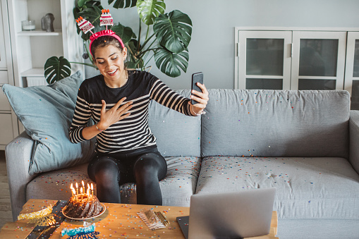 Teenage girl at home during pandemic isolation have birthday celebration and have video call with friends. Birthday cake with lighted candles on the table