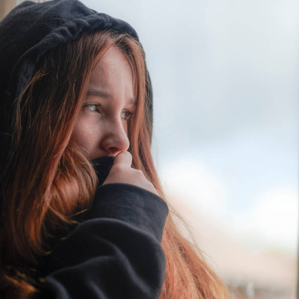 Gloomy female teenager depressed by secret violence standing by window and looking away while staying at home during pandemic of COVID 19 under isolation