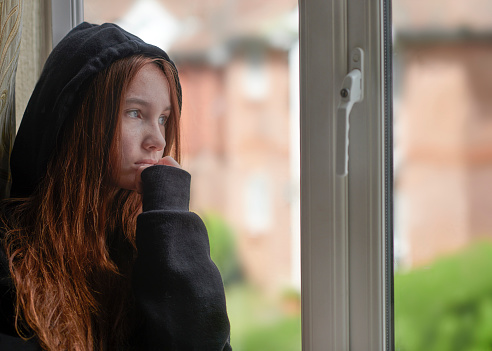 Side view of upset female teenager in black hoodie with red long hair depressed by secret abuse while standing next to window at home under isolation