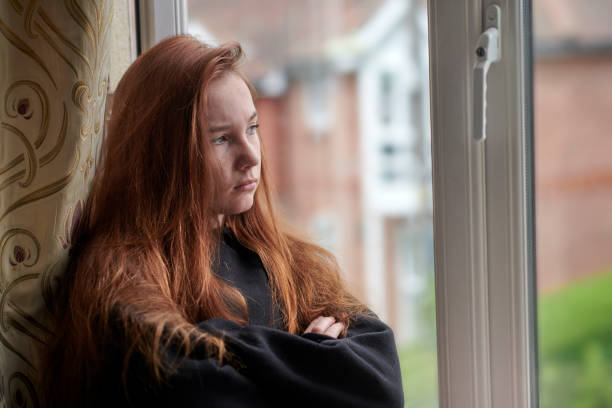 Upset teenage girl during quarantine at home looking out window Side view of moody female teenager in black casual wear with red hair standing at window with arms crossed and looking away while staying at home under isolation grumpy stock pictures, royalty-free photos & images