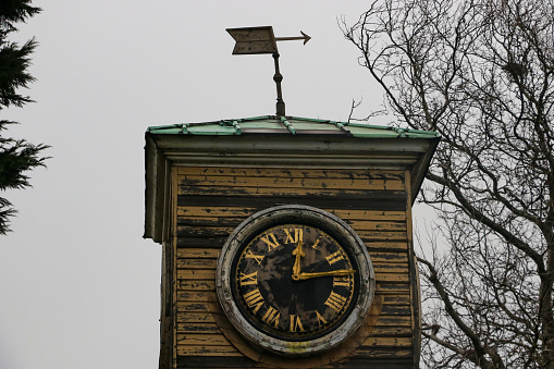 A late Gregorian period wooden clock tower with peeling paint and gold coloured clock hands and roman numerals, with a weather vain on its roof in winter in Sheerness dockyard, Kent, England, UK.