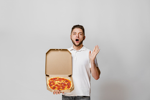 Surprised young bearded courier with pizza in cardboard box in hand. Crazy emotion. Stay at home and eat tasty pizza. Safety delivery without covid-19. Advertise for pizzeria at quarantine