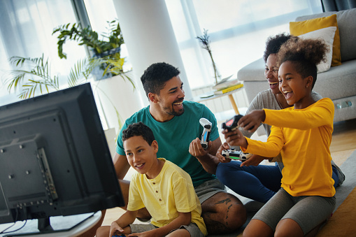 Closeup side view of a young african american family with two children spending time at home during coronavirus pandemic in 2020. They are playing some video games in the living room. 
For later use this is a quite useful family lifestyle content.