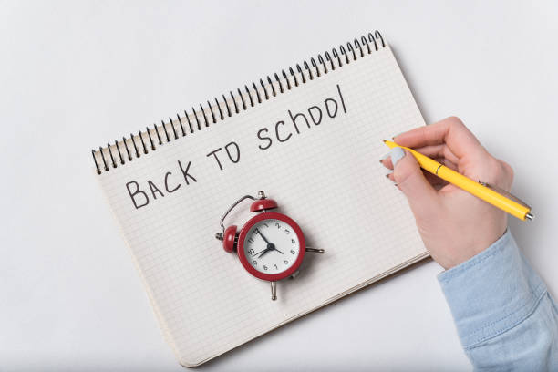 istock Female hand writes a phrase BACK TO SCHOOL. Vintage alarm clock. Top view 1223520979