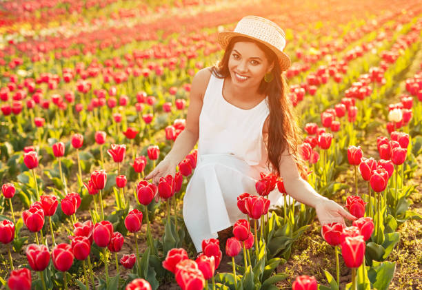 Optimistic woman in elegant dress and hat smiling and touching red tulips while sitting in meadow on sunny summer day