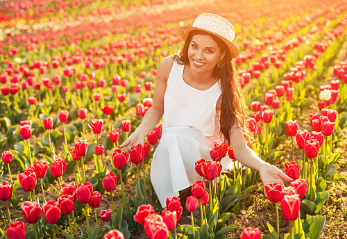 Optimistic woman in elegant dress and hat smiling and touching red tulips while sitting in meadow on sunny summer day