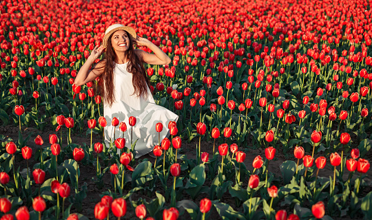 Optimistic elegant female adjusting hat and looking up while enjoying sunny day in red tulip field in spring