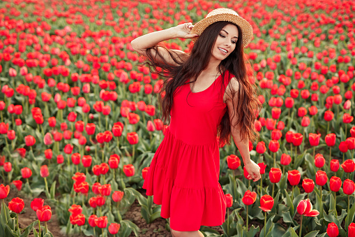 Charming carefree young long haired brunette in red sundress and straw hat enjoying nice day while standing on field among red tulips