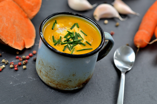 Sweet potato soup with cream and fresh chive in old enamel cup. Garlic, pumpkin and half pieced carrot in background. Slate slab and spoon.