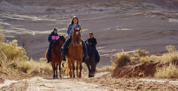 three native american (navajo) young people ride their horses with their pet dog in the arizona desert with a rock wall behind them on a bright, sunny day - monument valley navajo mesa monument valley tribal park stock-fotos und bilder