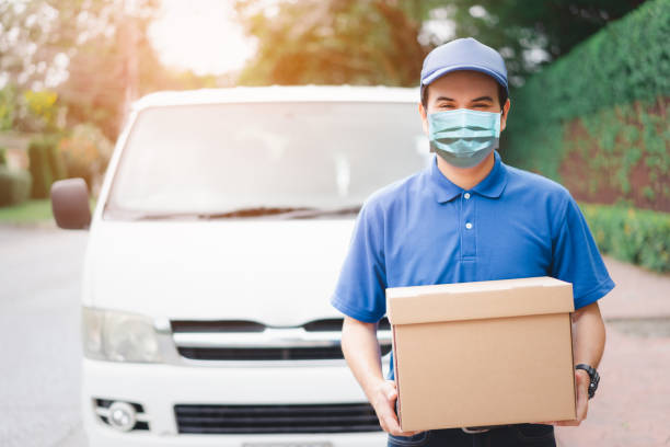 Postal delivery courier man wearing protective face mask in front of cargo van delivering package holding box due to Coronavirus disease or COVID-19 outbreak situation in all of landmass in the world. Postal delivery courier man wearing protective face mask in front of cargo van delivering package holding box due to Coronavirus disease or COVID-19 outbreak situation in all of landmass in the world. landmass stock pictures, royalty-free photos & images