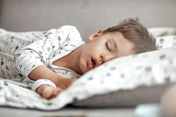 Cute little boy sleeping. Tired child taking a nap in parent's bed. Clean, fresh and cozy bedding sheets. Bedtime for kids. Sleepy toddler boy sleeping on his bed