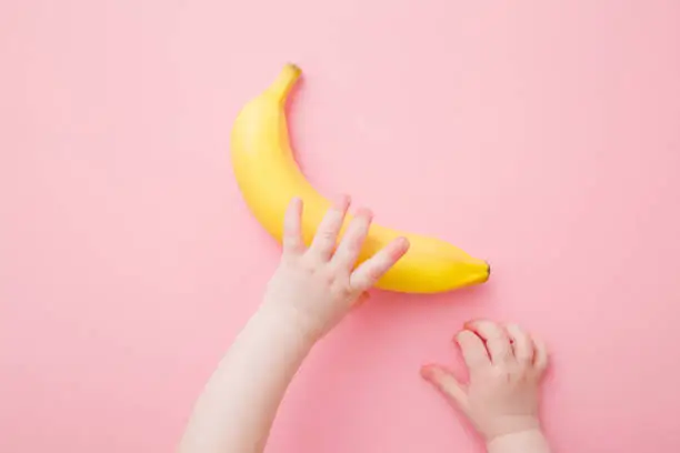 Baby hands taking yellow banana. Fresh fruit on light pink table background. Pastel color. Closeup. Point of view shot. Top down view.
