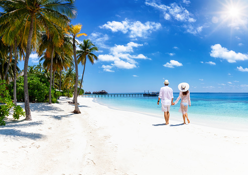 A holiday couple in white summer clothing walks down a paradise beach
