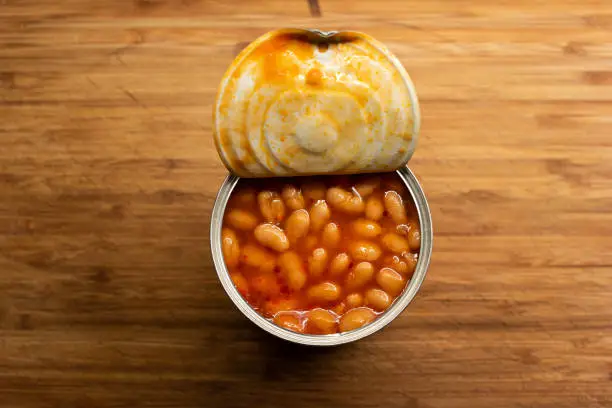 Opened tin can of generic store bough canned beans on a chopping wood board in the kitchen top view 2020