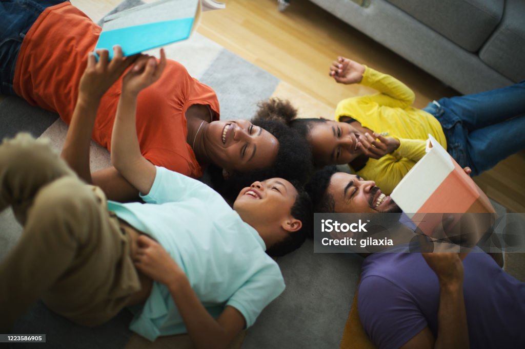 Home schooling is fun. Closeup top view of young african american couple home schooling their children. They are laying on the living room floor and parents are reading to the children during coronavirus quarantine. Adult Stock Photo