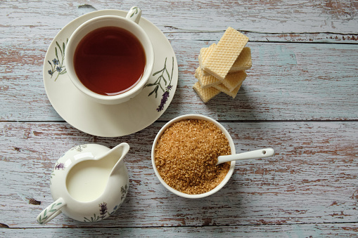 Top view of a mug with red tea, wafer biscuits, milk and brown cane sugar on wooden table. Flat lay