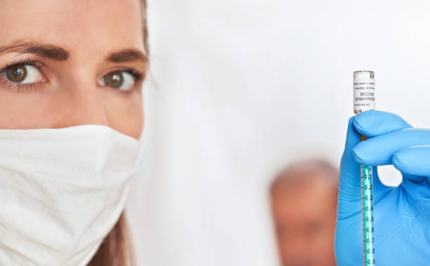 young nurse with white cotton mouth virus face mask, injecting coronavirus vaccine (sticker is own design) into green syringe, blurred unrecognisable patient in background. covid-19 cure concept - nitril imagens e fotografias de stock