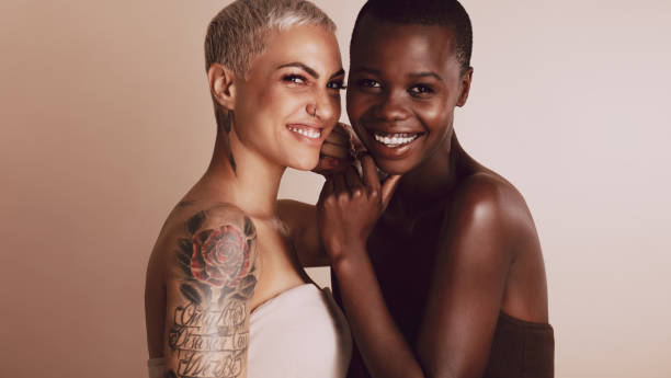Two beautiful women smiling together Two beautiful women standing together against beige background. Female models of different ethnicities with short hairstyle looking at camera and smiling. shaved black women stock pictures, royalty-free photos & images
