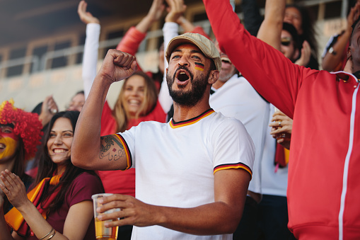 Crowd of spectators cheering at sports event, with a man holding a glass of beer. Germany football team supporters actively chanting in stadium.