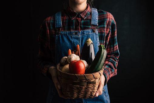 A woman wearing overalls and a checked shirt holding a basket full of vegetables on a black background.